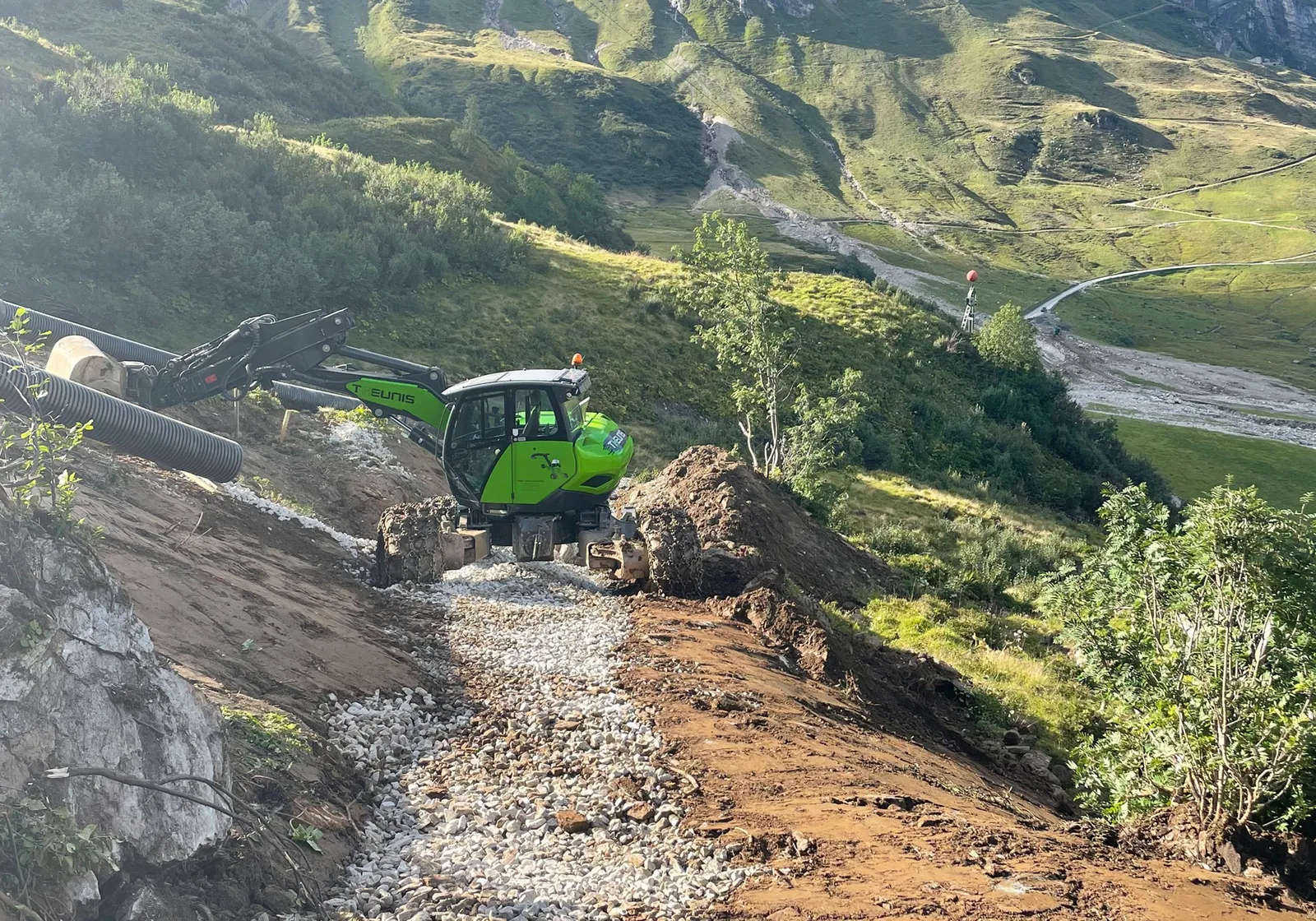 Grüner Schreitbagger baut einen geschotterten Weg an einem steilen Berghang